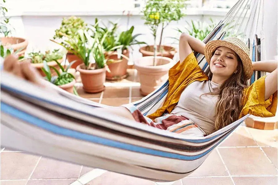 Une femme portant un chapeau de paille se détend dans un hamac rayé entouré de plantes en pot.